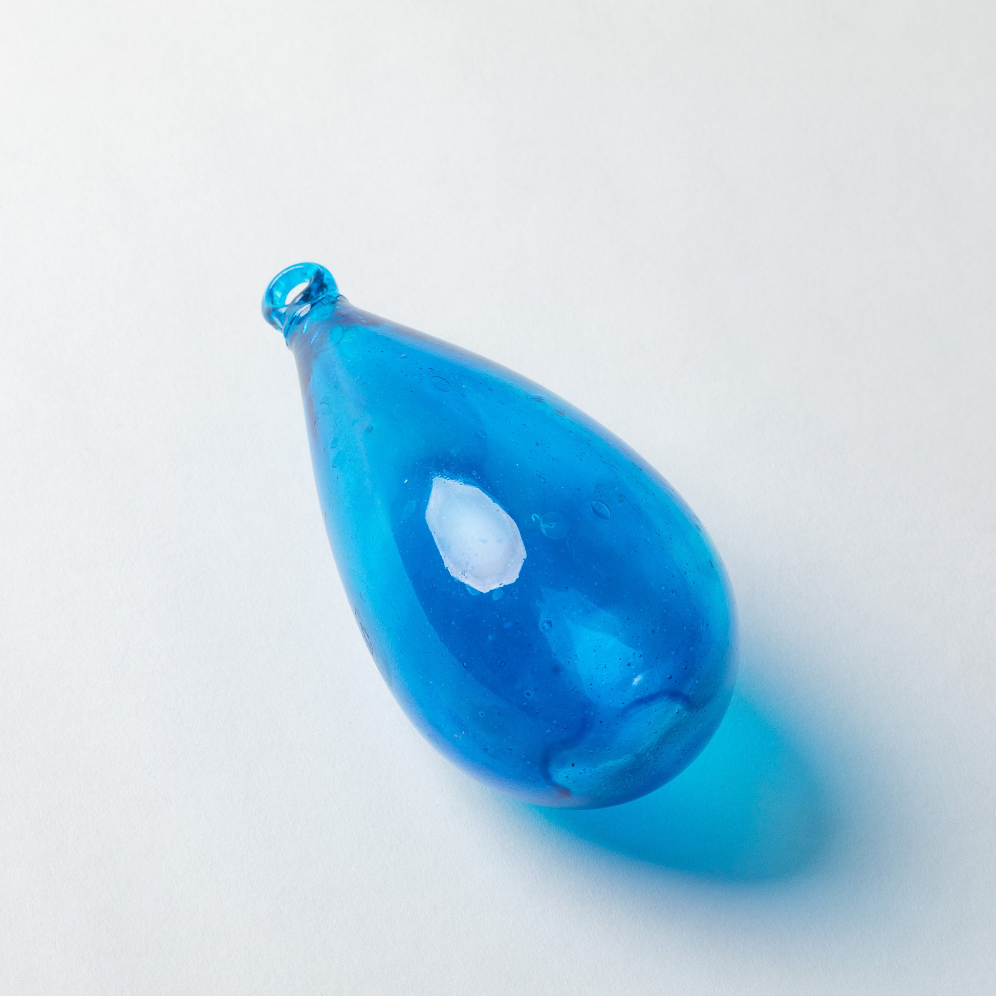 A translucent blue glass teardrop pendant with a small bubble-filled neck, resting on a light gray background with a soft blue shadow.