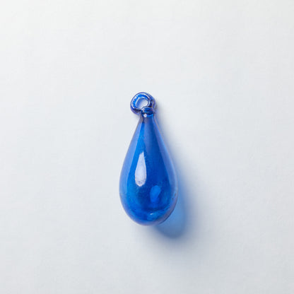 A glossy, teardrop-shaped blue glass pendant with a small loop at the top, shown on a clean, light gray background.