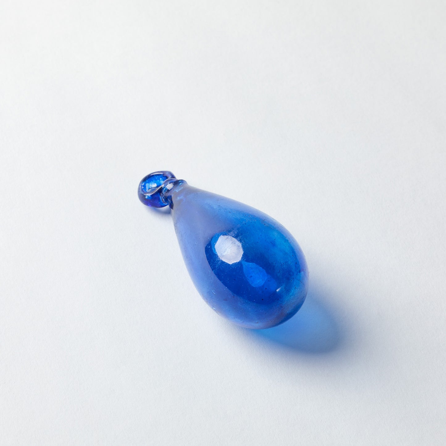 A teardrop-shaped blue glass pendant with a small faceted blue top, photographed on a clean white background with a soft shadow.