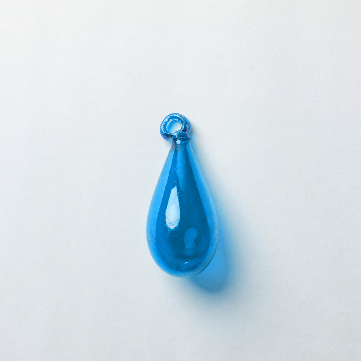 A translucent blue teardrop-shaped glass pendant with a small loop at the top, shown on a plain light gray background.
