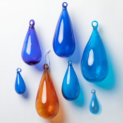 Seven teardrop-shaped glass ornaments in various shades of blue and one amber-orange, arranged on a clean white background.