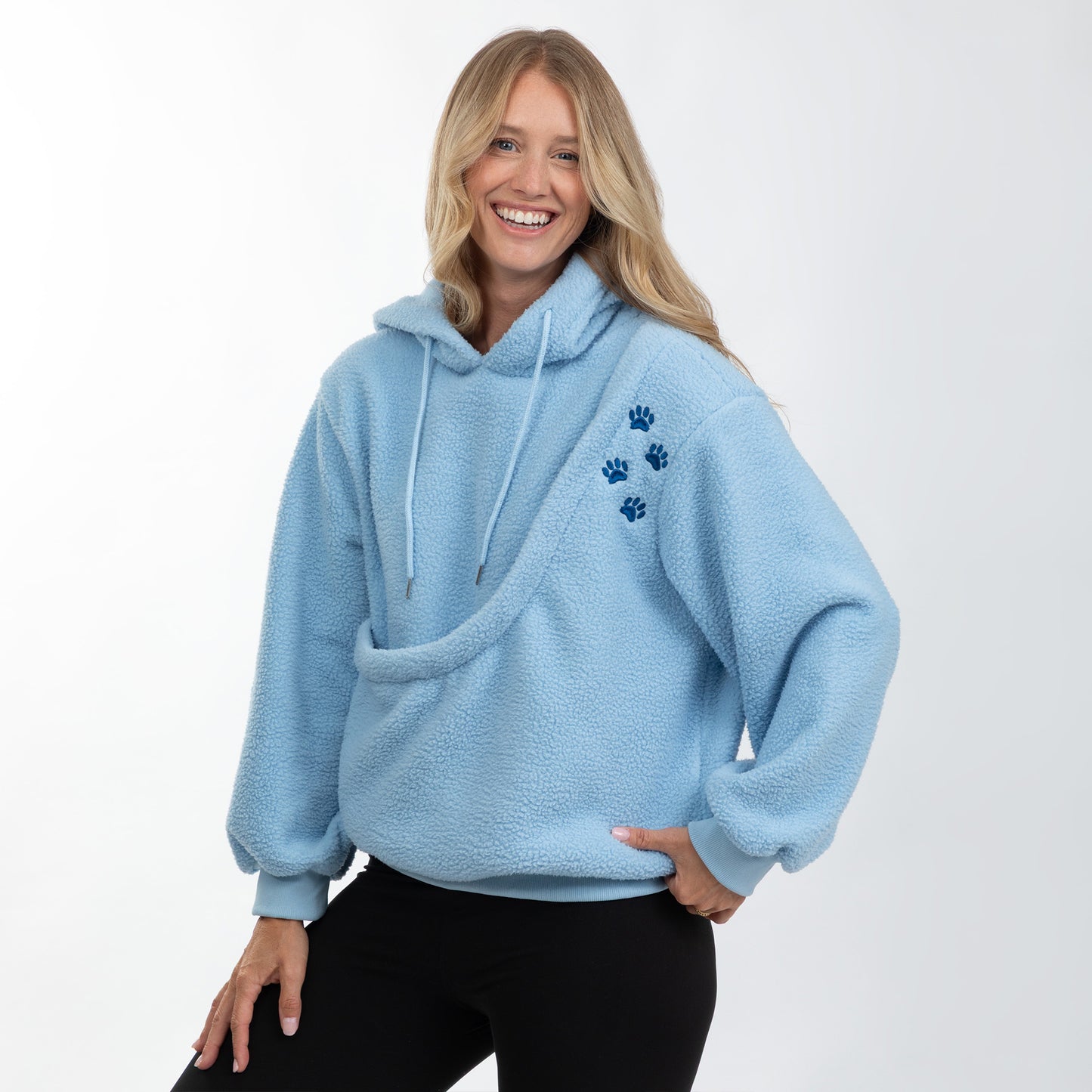 A smiling blonde woman wearing a light blue sherpa fleece hoodie with three dark blue paw prints on the left chest, paired with black pants, against a plain white studio background.