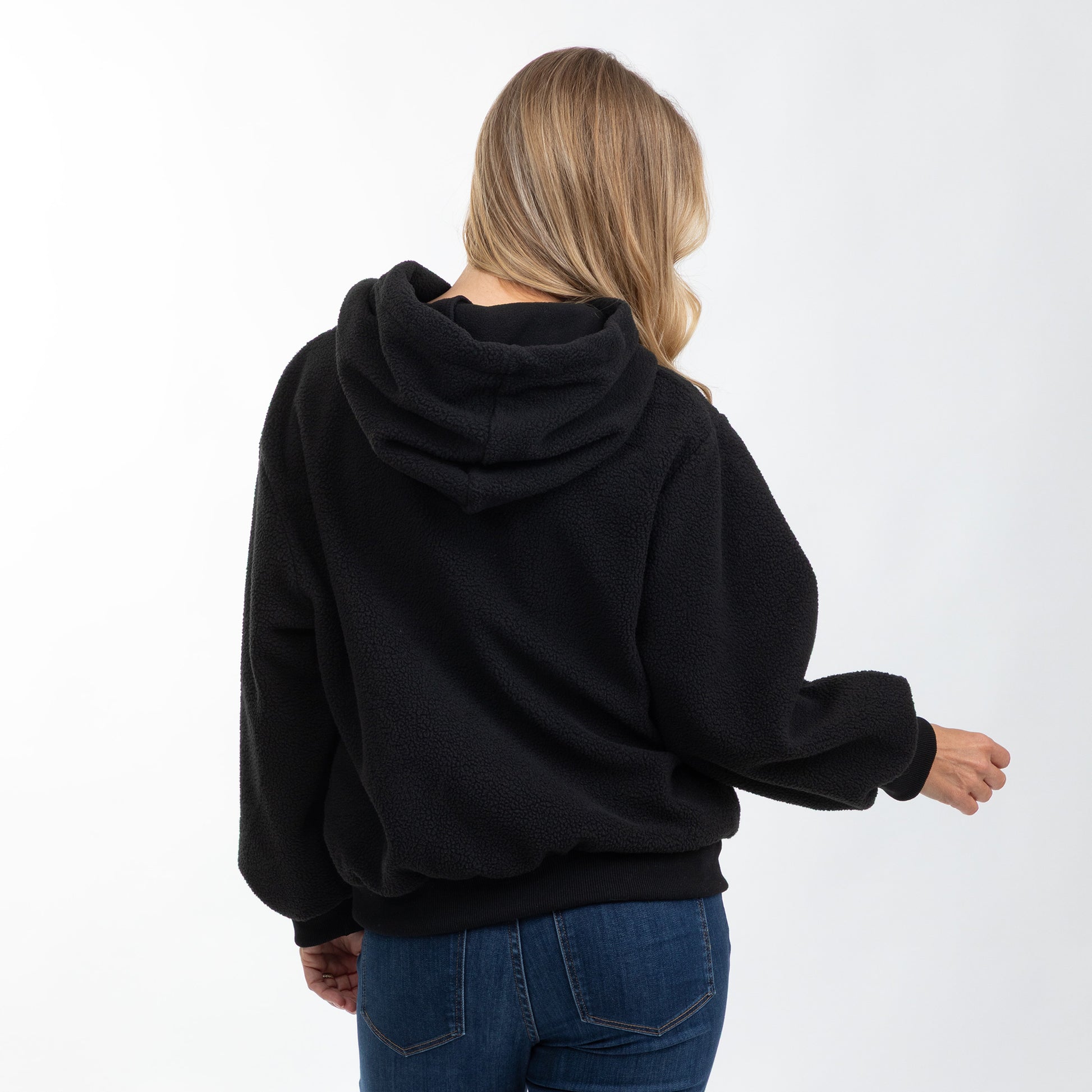A woman viewed from behind wearing a black fleece pullover hoodie and blue jeans against a plain white studio background.