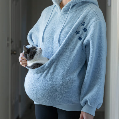 A person wearing a light blue sherpa hoodie with blue paw prints holds a small gray and white cat indoors.