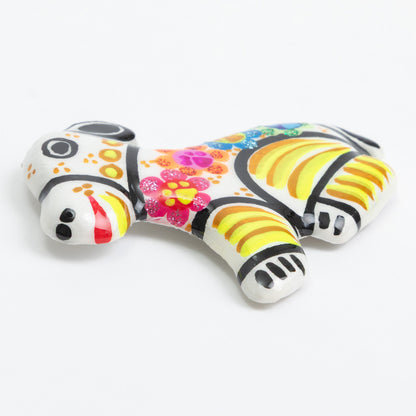 A small decorative magnet shaped like a dog, painted with a white base and colorful floral and striped patterns in pink, yellow, orange, blue, and black, photographed on a clean white background.
