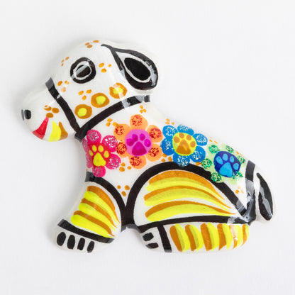 A small decorative magnet shaped like a dog, painted with a white base and colorful floral and striped patterns in pink, yellow, orange, blue, and black, photographed on a clean white background.