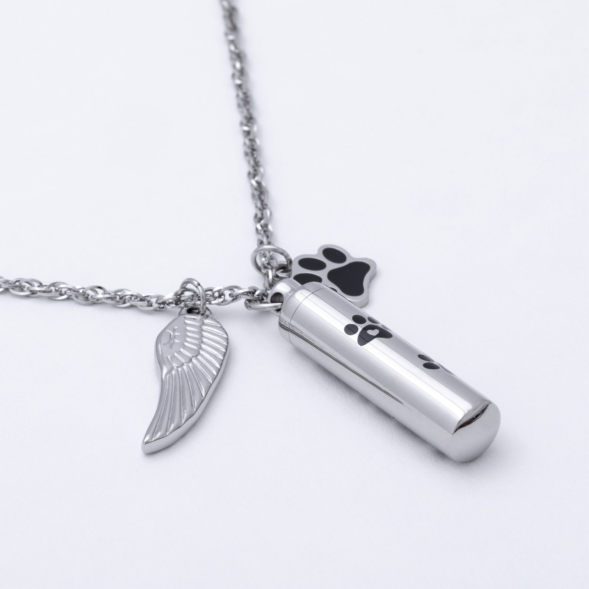 Silver-toned metal cremation urn necklace with a cylindrical urn pendant, a wing-shaped charm, and a paw print charm, shown on a white background.
