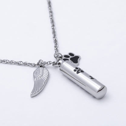 Silver-toned metal cremation urn necklace with a cylindrical urn pendant, a wing-shaped charm, and a paw print charm, shown on a white background.