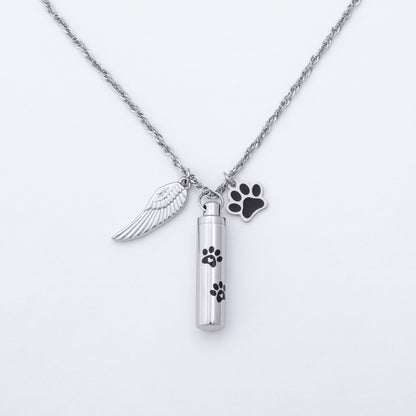 A silver-tone cremation urn necklace featuring a cylindrical vial pendant with black paw print engravings, an angel wing charm, and a black paw-shaped tag on a white background.