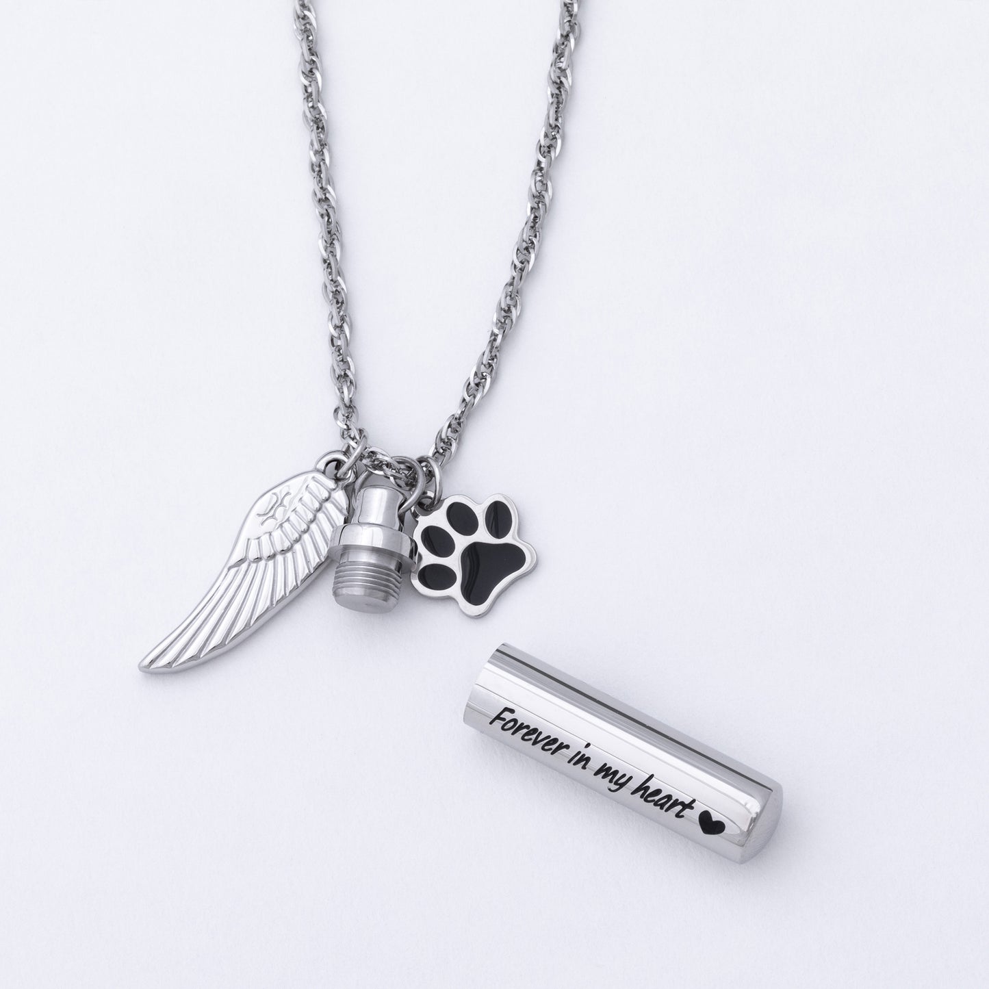 Silver-tone memorial necklace with a wing charm, a paw print charm, a small urn charm, and a cylindrical keepsake tube engraved with 'Forever in my heart' on a white background.