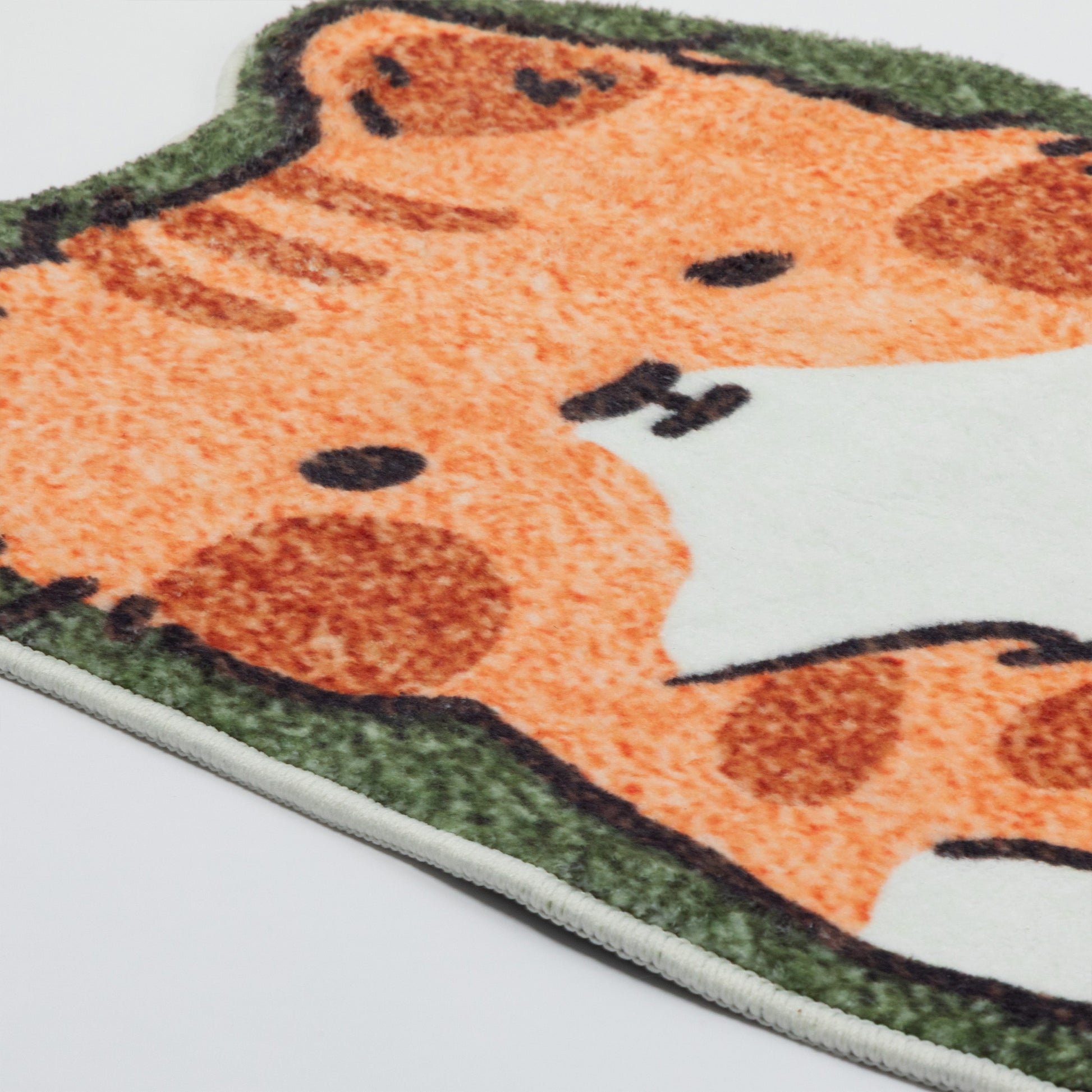 A close-up of a plush rug featuring a cartoon cat design in orange, brown, and white, with a green textured border, shown on a white background.