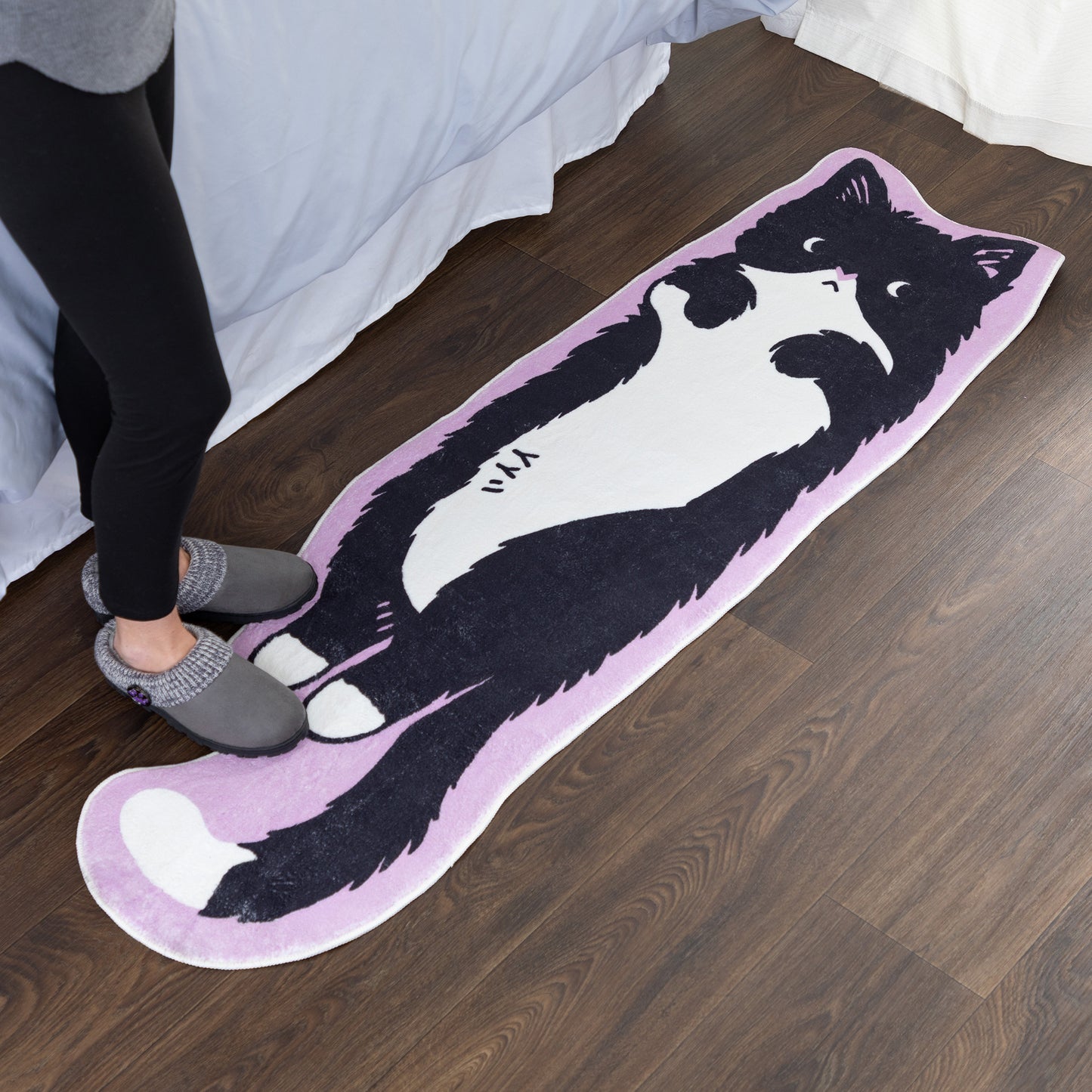 A long, cat-shaped floor mat with a black and white cat illustration on a light purple background, shown on a dark wood floor next to a bed with a person's legs and slippers visible.