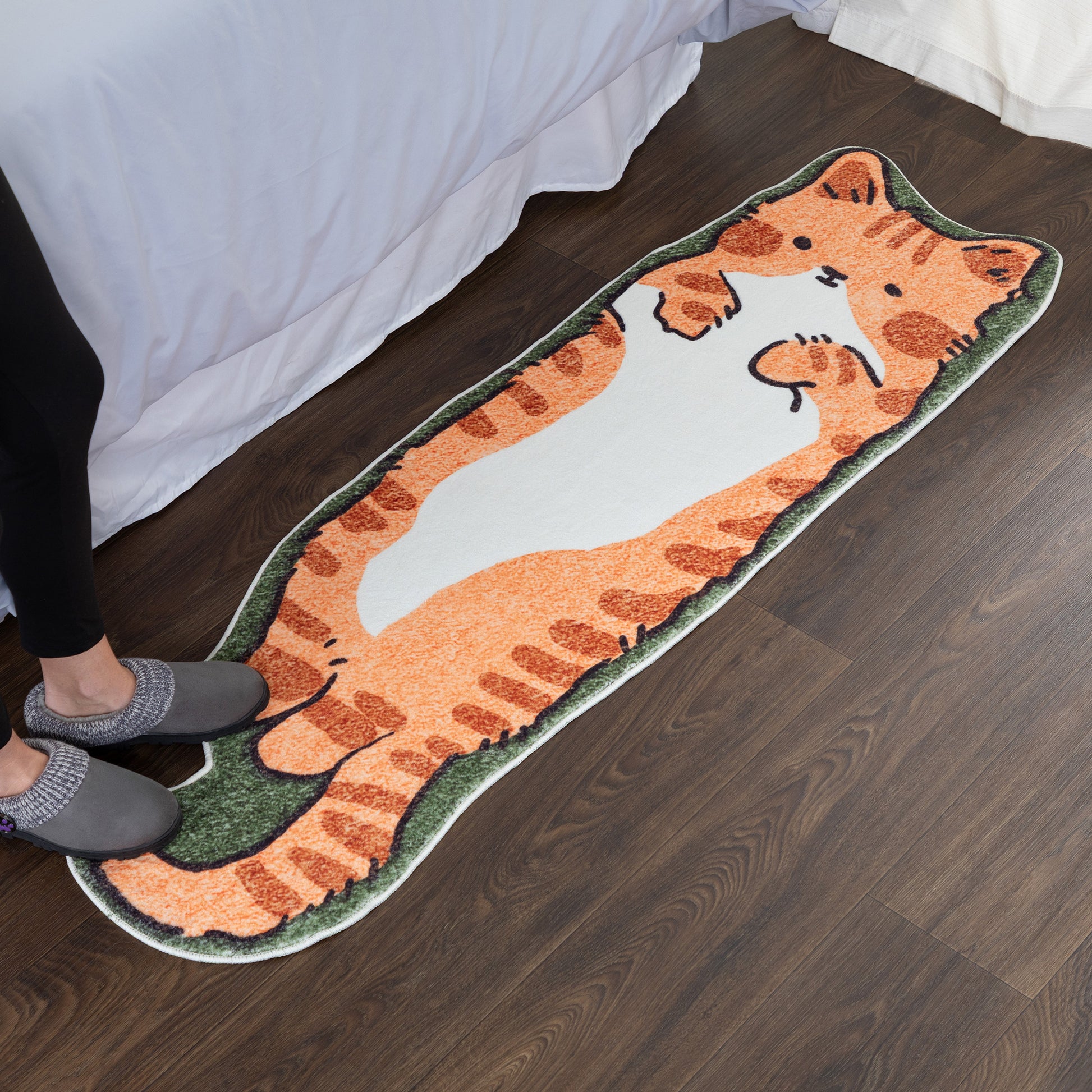 A long, cat-shaped rug with a cartoon cat illustration lies on a dark wood floor next to a bed. A person's legs and feet wearing gray slippers are visible at the bottom left, indicating a lifestyle setting.