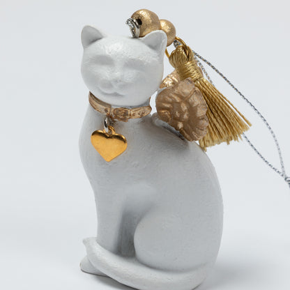 A white cat figurine wearing a gold collar with a gold heart-shaped tag, a gold tassel ornament, and a small gold bell, set against a plain light gray background.