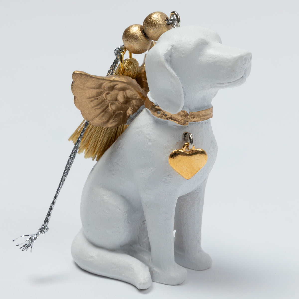 A white dog figurine styled as an angel, featuring gold metallic wings, a gold heart-shaped tag, and gold tassels on a silver chain, set against a plain light gray background.