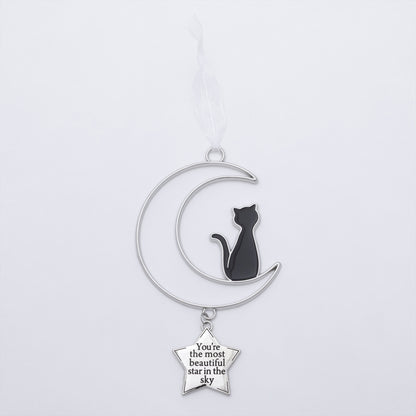 A silver crescent moon and star ornament hanging from a white ribbon. The crescent moon features a black enamel cat silhouette, and a small silver star charm below reads, 'You're the most beautiful star in the sky'.