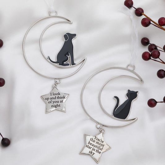 Two silver crescent moon and star ornaments displayed on a white fabric background. One ornament features a black dog silhouette on the crescent moon with a dangling silver star that reads 'I look up and think of you at night.