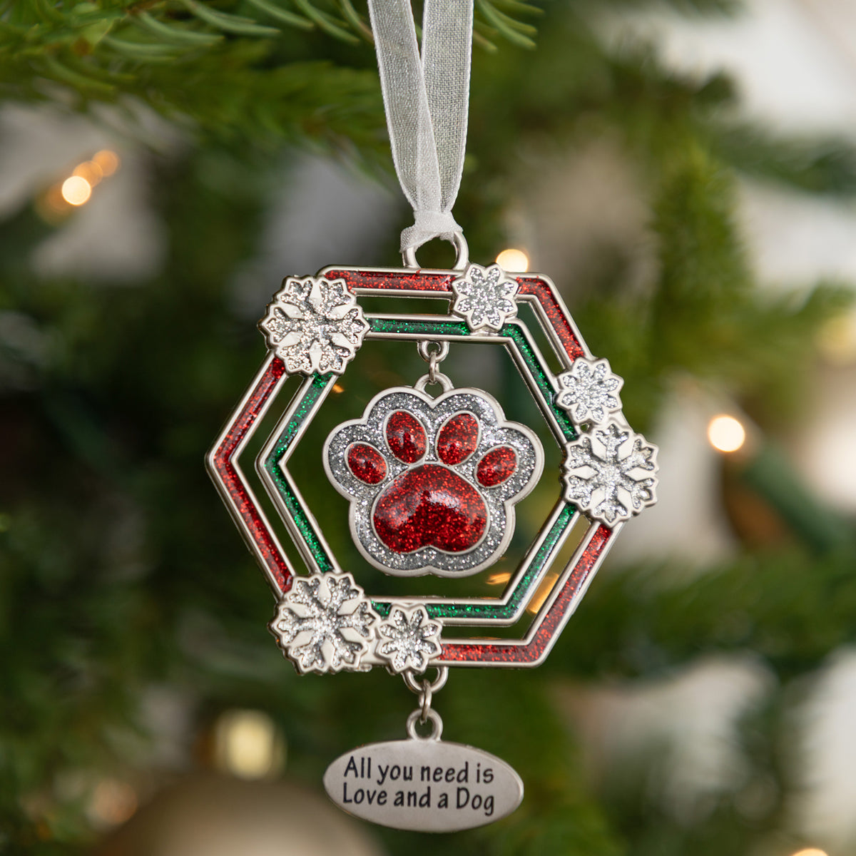A silver-toned dog paw print Christmas ornament hanging from a white ribbon, featuring red glitter paw print details, a red and green hexagonal frame, and a dangling silver tag that reads 'All you need is Love and a Dog' against a blurred Christmas tree background.