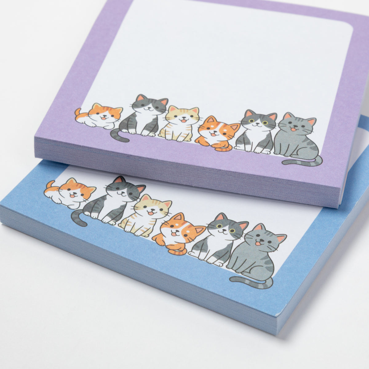 Two pastel notepad sheets stacked on a white background, each printed with a row of cute illustrated cats. The top pad has a lavender border and the bottom pad has a light blue border.