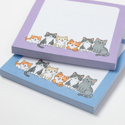 Two pastel notepad sheets stacked on a white background, each printed with a row of cute illustrated cats. The top pad has a lavender border and the bottom pad has a light blue border.