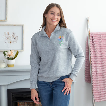 A woman wearing a light gray quarter-zip sweatshirt with a colorful floral design on the left chest, paired with blue jeans, standing in a bright, modern room.