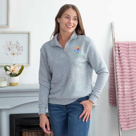 A woman wearing a light gray quarter-zip sweatshirt with a colorful floral design on the left chest, paired with blue jeans, standing in a bright, modern room.