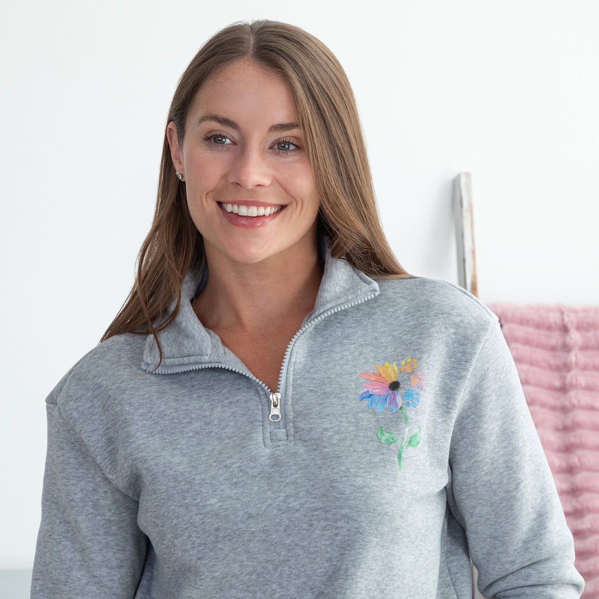 A smiling woman wearing a heather gray quarter-zip pullover sweatshirt with a colorful flower on the left chest, set against a bright white indoor background.
