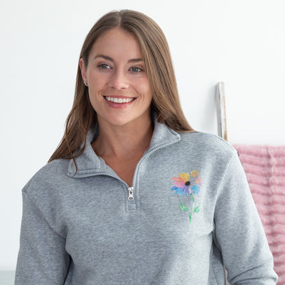 A smiling woman wearing a heather gray quarter-zip pullover sweatshirt with a colorful flower on the left chest, set against a bright white indoor background.
