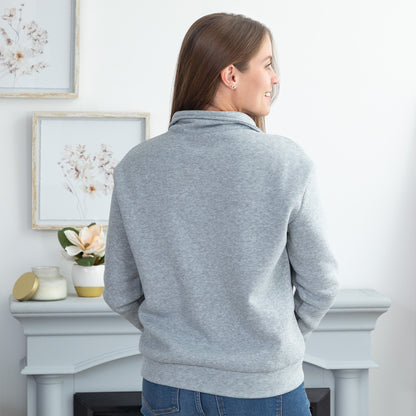 A woman viewed from the back wearing a light gray heather mock-neck sweatshirt, standing in front of a white fireplace mantel in a bright room.