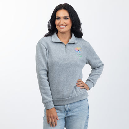 A woman modeling a light gray heathered quarter-zip sweatshirt with a small colorful embroidered flower on the left chest, paired with light blue jeans, against a plain white studio background.