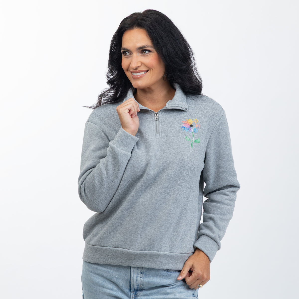 A woman wearing a heather gray quarter-zip sweatshirt with a colorful embroidered flower on the left chest, paired with light blue jeans, against a plain white background.
