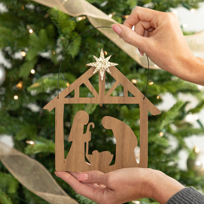 A wooden nativity scene decor held by two hands in front of a decorated Christmas tree. The ornament features a light brown wood cutout of Mary, Joseph, and baby Jesus in a stable, with a glittery gold star on top and a black hanging loop.