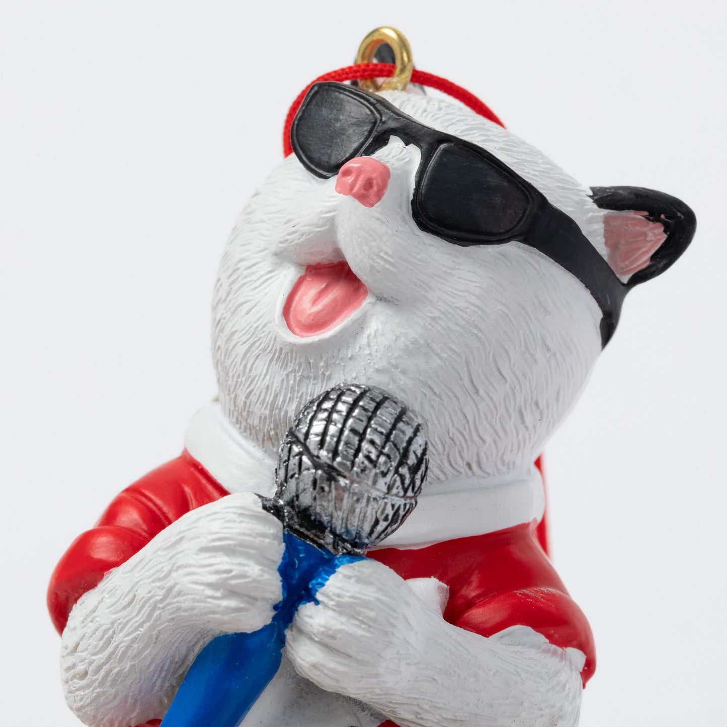 A white cat ornament wearing black sunglasses, a red Santa-style jacket, and a blue bow tie, holding a silver microphone and singing into it. The figurine has a gold hanging loop at the top and is photographed against a plain light gray background.