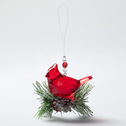 A red plastic cardinal Christmas ornament perched on green pine branches with a small pinecone, hanging from a clear string against a light gray background.