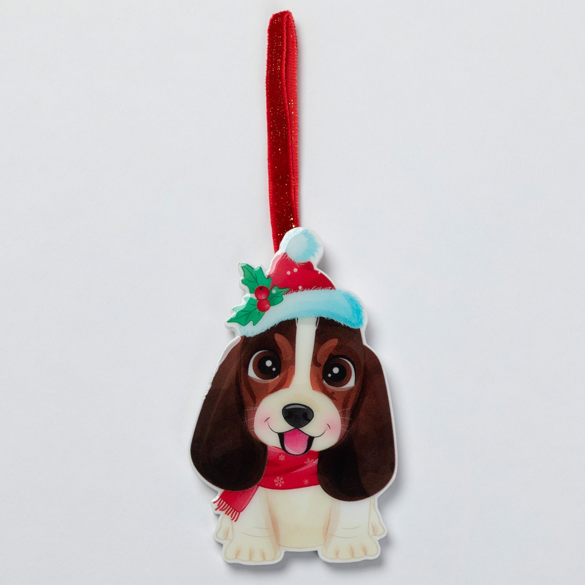 A Christmas dog ornament featuring a brown and cream Bichon Frise illustration wearing a red Santa hat with green holly and a red scarf, hanging from a red glitter ribbon on a light gray background.