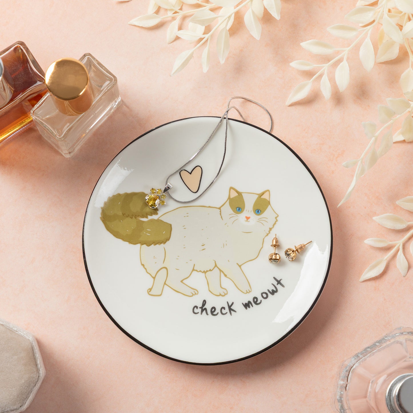 A round white ceramic jewelry tray with a black rim, decorated with a cute illustration of a white cat with green ears and the text 'check meout'. The tray is styled on a pink background with decorative dried leaves, perfume bottles, and small gold earrings.