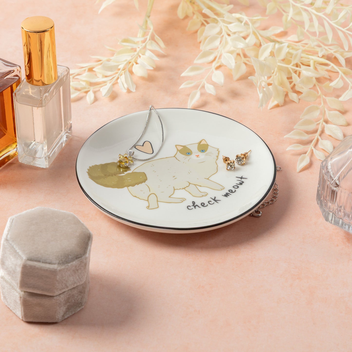 A round white jewelry tray with a black rim and a cat illustration, styled on a peach background with perfume bottles, a gray velvet box, dried flowers, and small gold earrings.