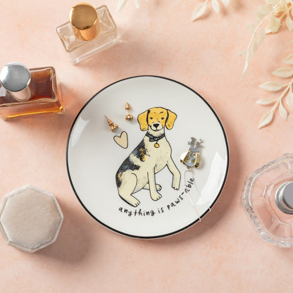 A round white ceramic jewelry tray with a black rim, featuring a cartoon beagle illustration and the text 'anything is paws-table'. The tray is styled on a peach-pink surface with perfume bottles, a small jewelry box, and decorative leaves around it.