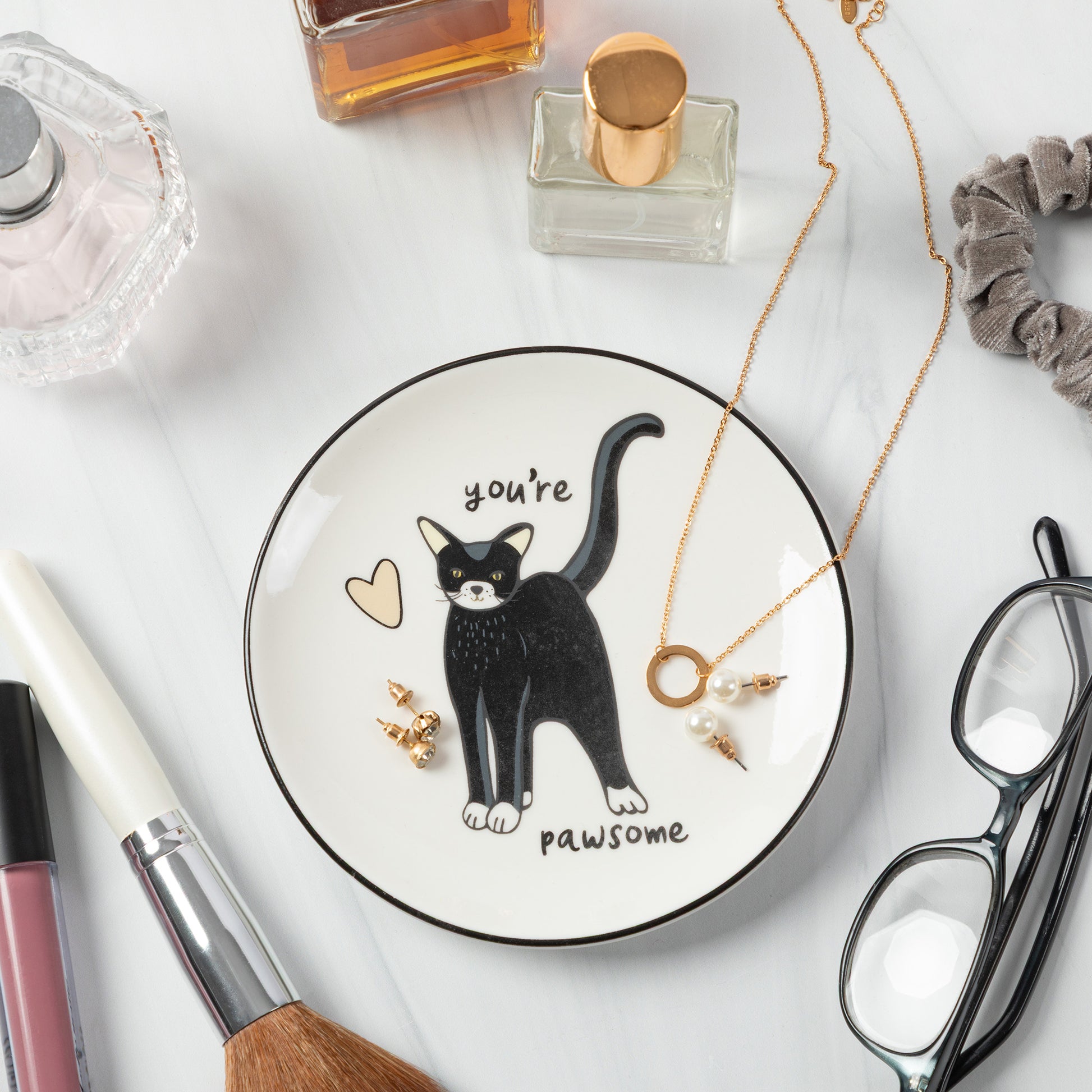 A round white jewelry tray with a black rim, featuring a black cat illustration and the words 'you're pawsome'. The tray is styled on a white marble surface with various cosmetic and eyewear props.