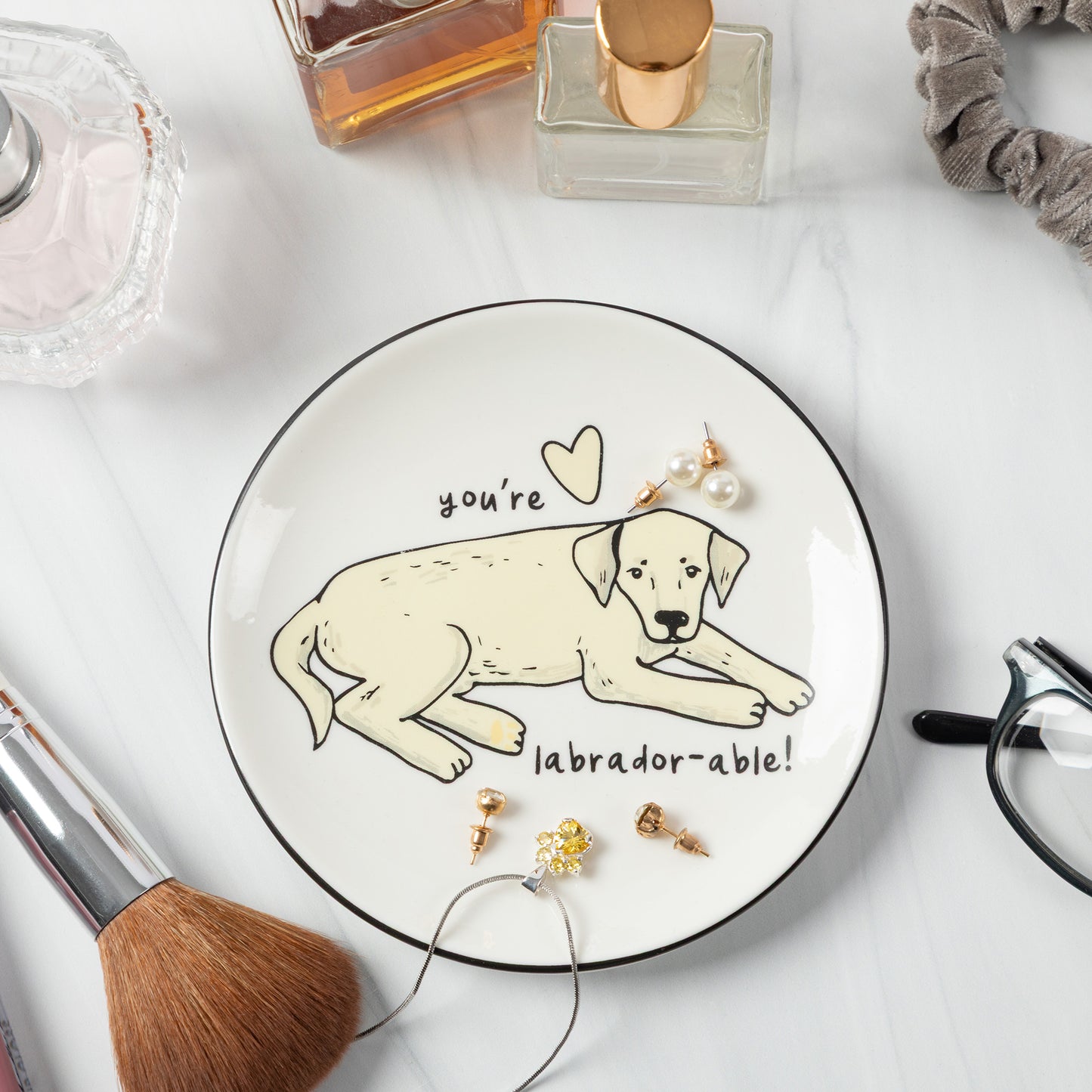 A round white ceramic plate with a black rim, featuring a cartoon Labrador retriever and the text 'you're labrador-able!' in black. The plate is styled in a flat-lay arrangement with various beauty and lifestyle props on a white marble surface.