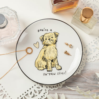 A round white ceramic jewelry tray with a black rim, featuring a yellow sketch of a small dog and the text 'you're a su-paw star!'. Gold-tone jewelry pieces and a necklace are arranged on and around the tray, which sits on a white lace doily over a light wood surface with perfume bottles in the background.