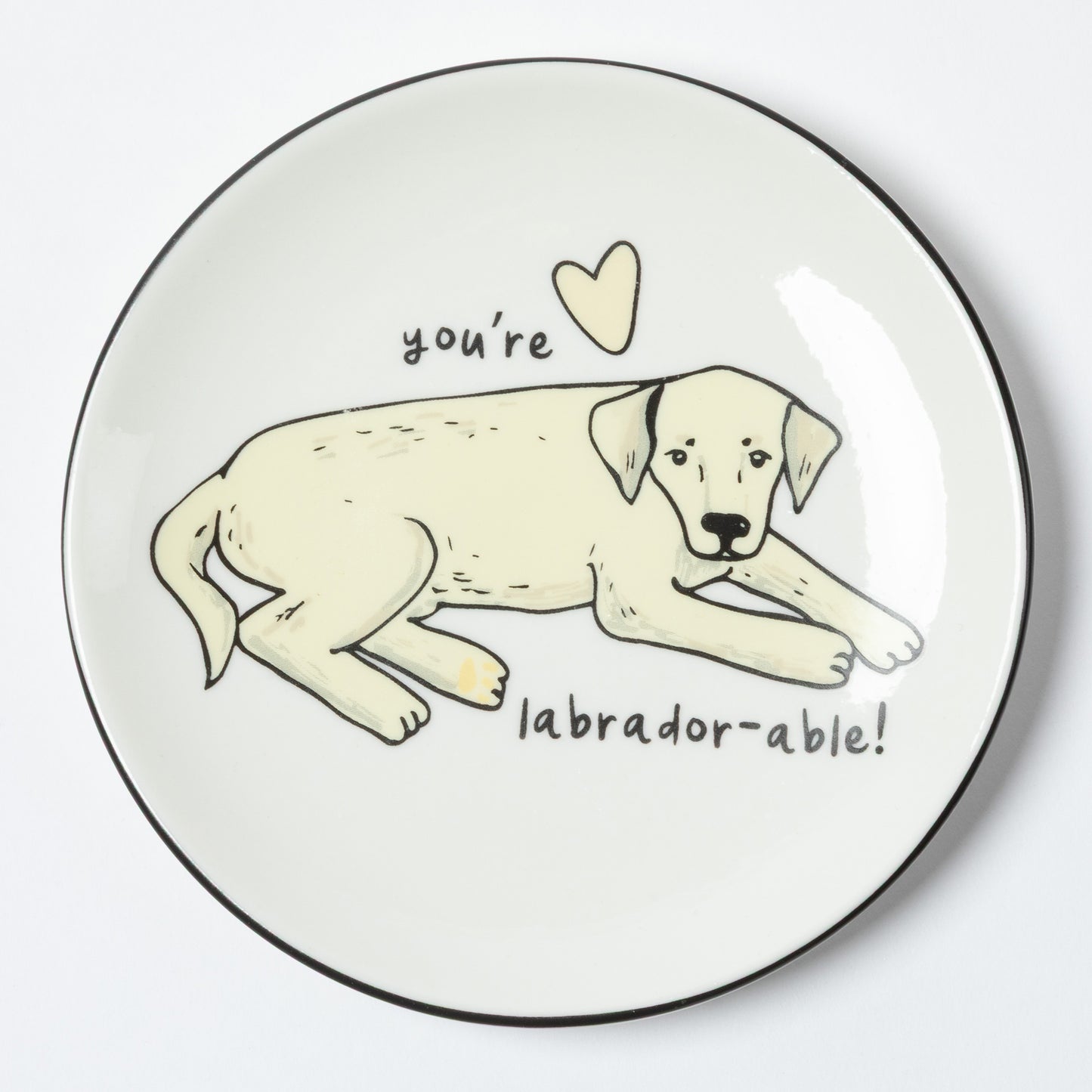 A round white plate with a black rim, featuring a hand-drawn illustration of a light cream Labrador puppy lying down with the text 'you're labrador-able!' and a small heart above it.