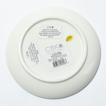 The underside of a white ceramic plate or shallow bowl, showing printed labels and a barcode sticker. The brand name CBK is clearly visible on the labels.