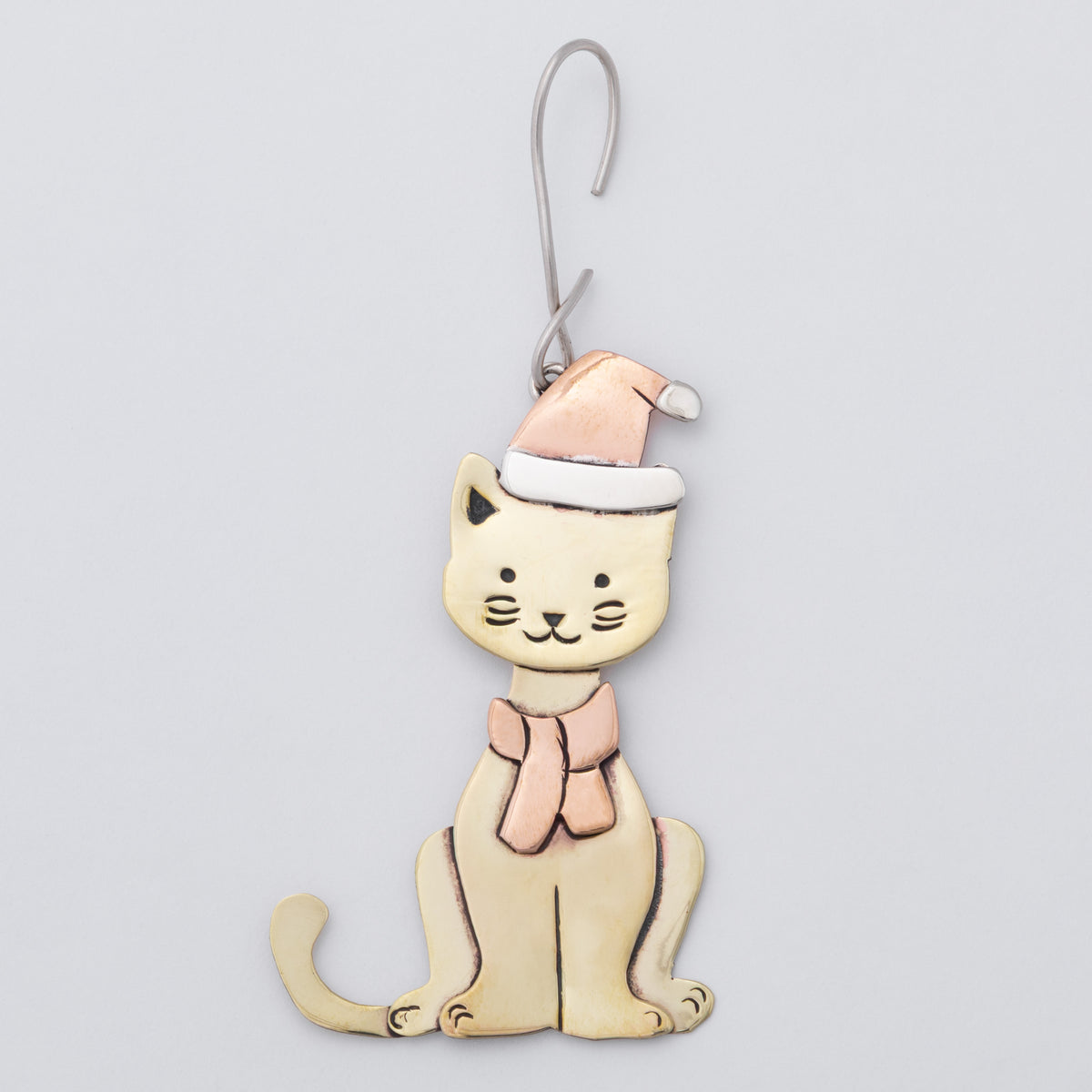 A whimsical cat-shaped ornament with brass body, a copper Santa hat, and matching scarf, hanging from a silver metal hook against a light gray background.