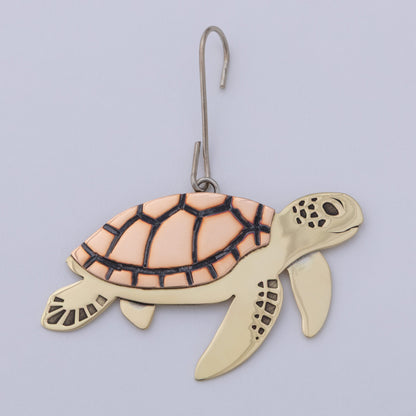 A sea turtle ornament with a gold-toned body and a peach-colored shell outlined in black, hanging from a silver metal hook against a light gray background.
