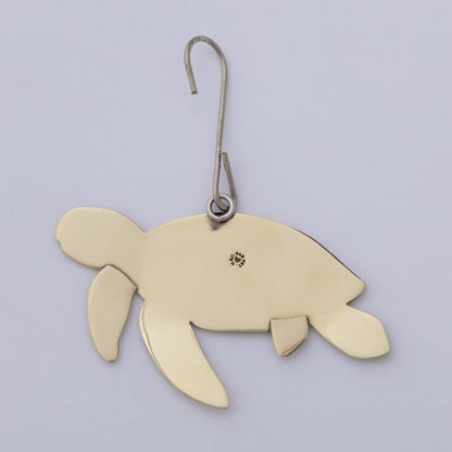 A sea turtle-shaped metal ornament in a warm gold finish, suspended from a silver-colored metal hook with a small decorative emblem on its shell, set against a plain light gray background.