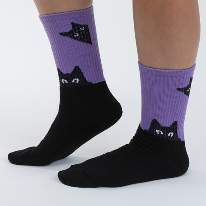 A pair of crew socks worn on a person's feet, featuring a black cat face and body design over a purple ribbed cuff, shown against a plain light gray background.