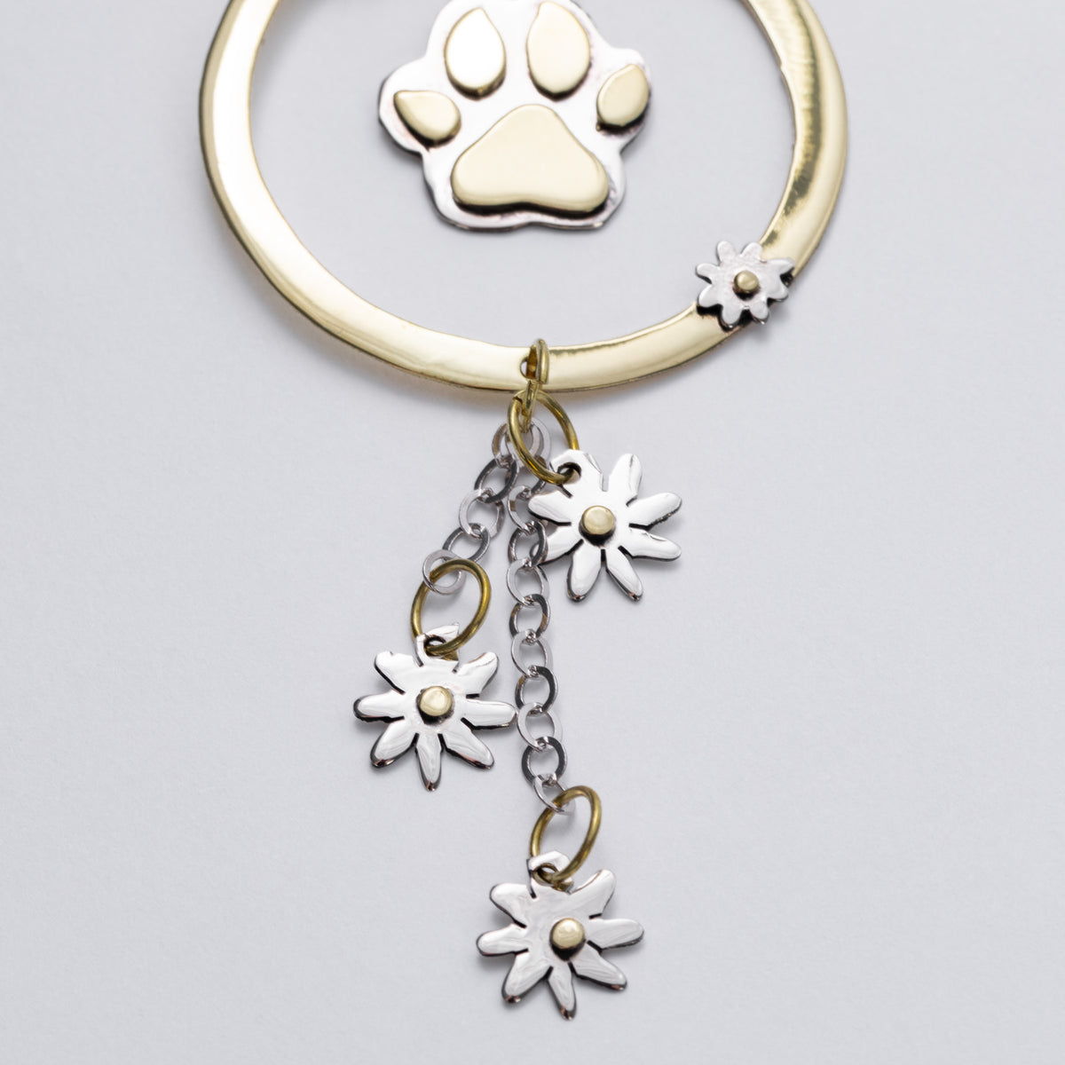 A gold and silver car charm featuring a paw print charm, a small flower accent, and three dangling daisy charms on a light gray background.