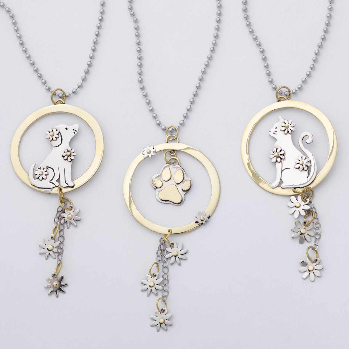 Three gold and silver pet memorial necklaces with circular pendants featuring dog, cat, and paw print designs, shown on a light gray background.