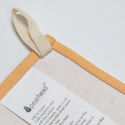 A close-up of a Pearhead cotton kitchen towel with a cream-colored loop handle and a warm orange border, featuring printed care instructions on a white paper insert.